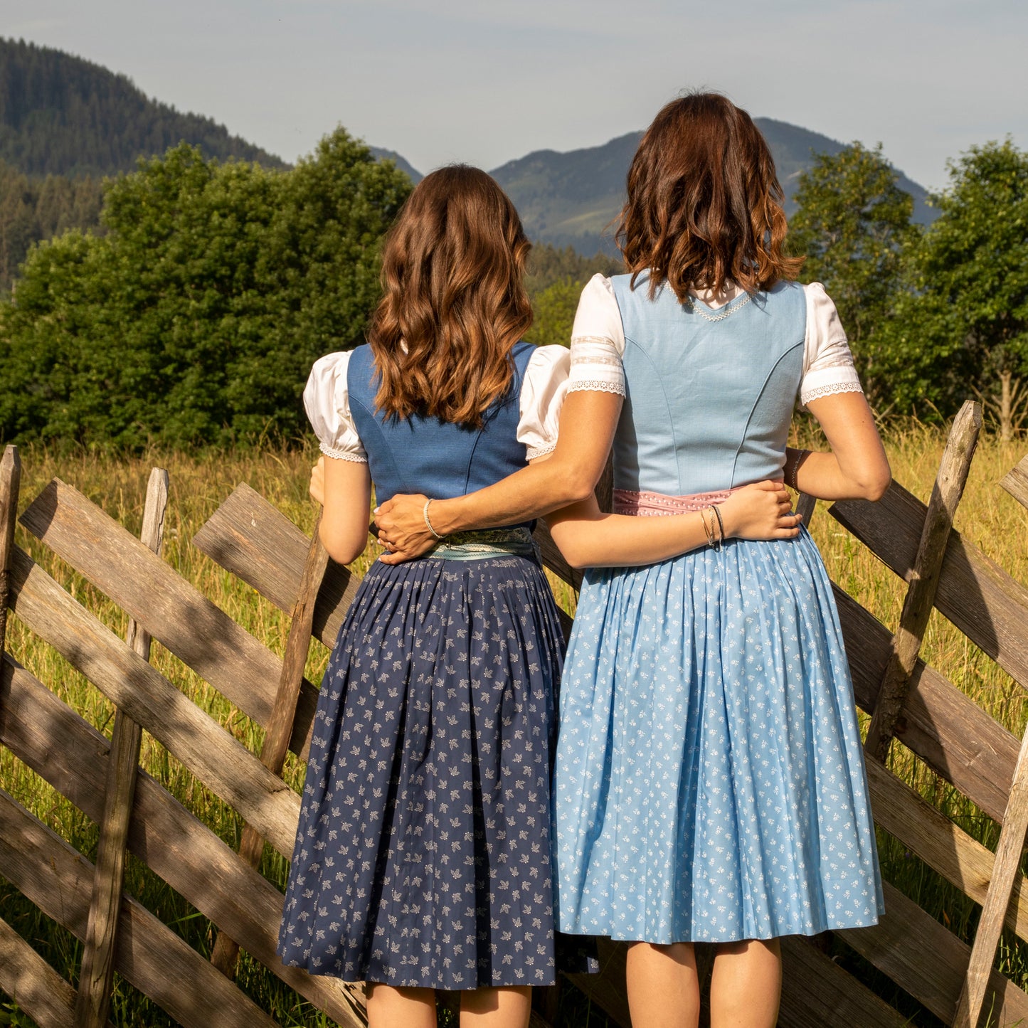 Zwei Frauen in Dirndl nebeneinander, von hinten, vor einem Holzzaun im Freien